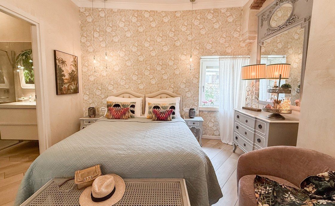 Schlafzimmer in Santo Stefano mit Blumentapete, salbeigrüner Tagesdecke und einem Strohhut auf der Bank